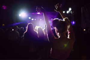 A dark venue with a girl with her hands up dancing to a band playing.